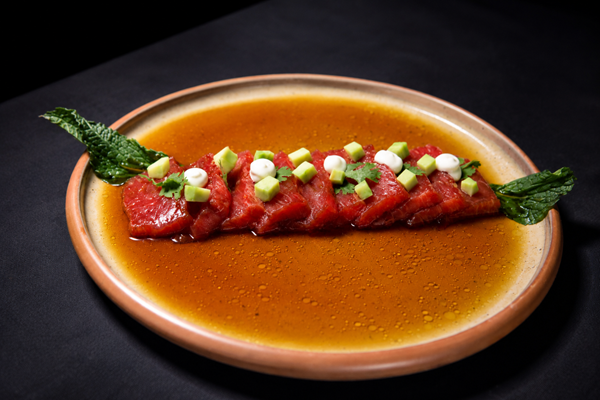 Tataki de sandía presentado en finas lonjas alineadas sobre una salsa oscura brillante, decorado con dados de aguacate, puntos de crema blanca y hojas de menta en un plato hondo de cerámica.