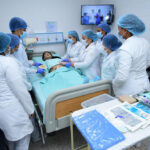 Siete estudiantes de salud vistiendo uniformes médicos, mascarillas y cofias quirúrgicas azules observan y realizan procedimientos de curación sobre un maniquí en un aula de simulación clínica, rodeados de insumos médicos e instrumentos sobre una mesa.