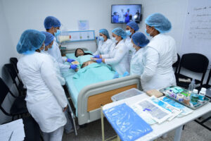 Siete estudiantes de salud vistiendo uniformes médicos, mascarillas y cofias quirúrgicas azules observan y realizan procedimientos de curación sobre un maniquí en un aula de simulación clínica, rodeados de insumos médicos e instrumentos sobre una mesa.